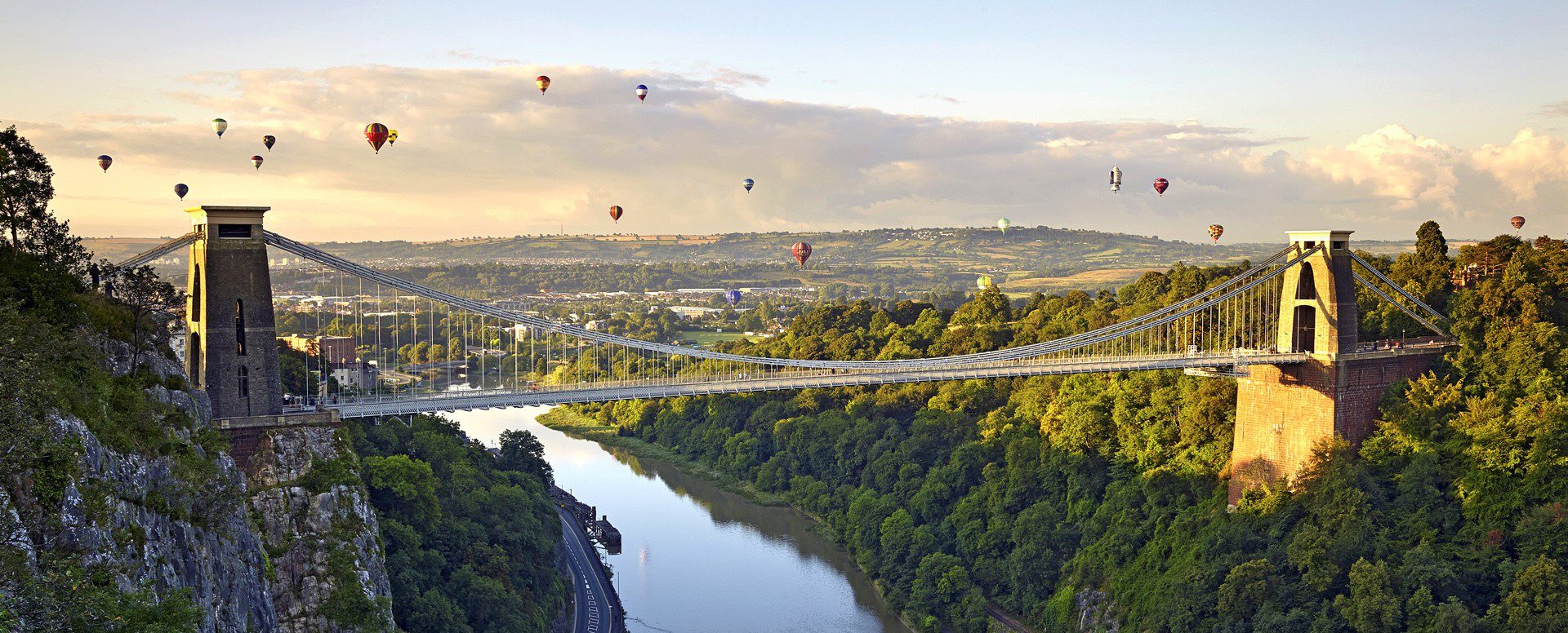 Picturesque shot of Clifton Suspension Bridge lit by the sun with around 15 hot air-balloons in the distance.